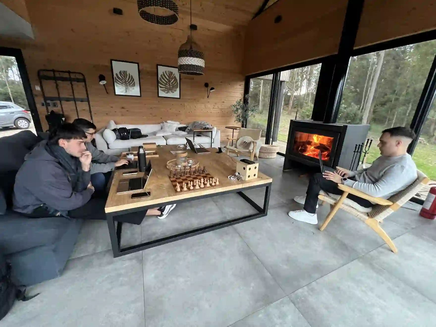Tres personas del equipo trabajando con notebooks alrededor de una mesa, en una casa con chimenea encendida y vista al bosque.